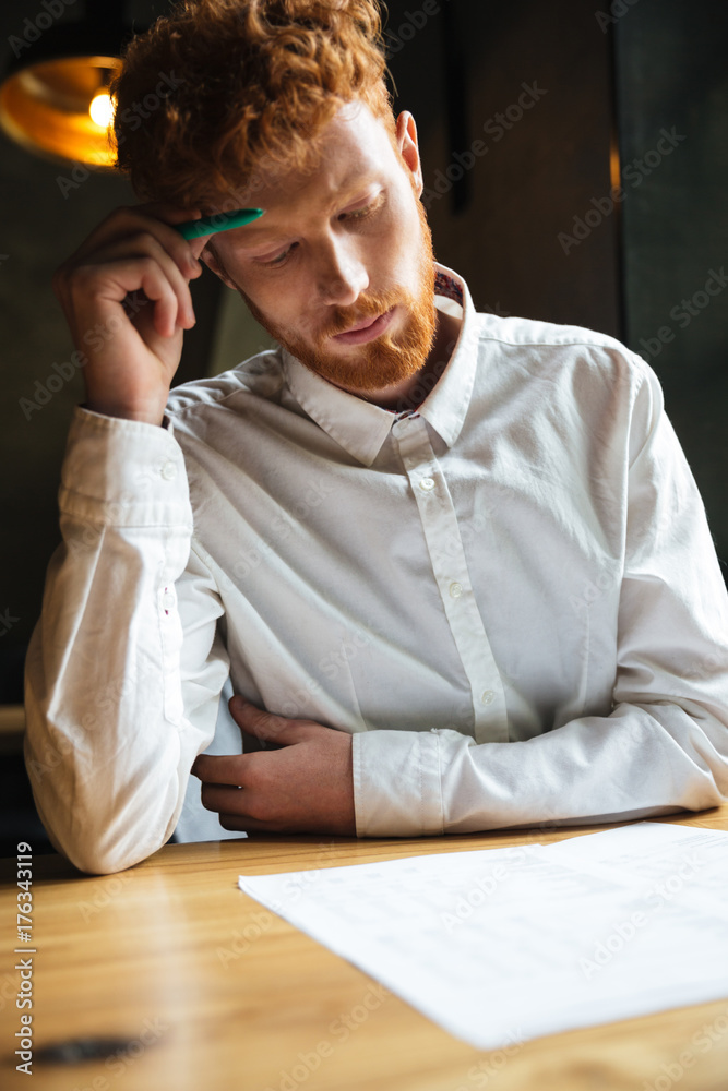 Portrait of concentrated readhead curly bearded man, reading papers , sitting at wooden table Portrait of concentrated readhead curly bearded man, reading papers , sitting at wooden table