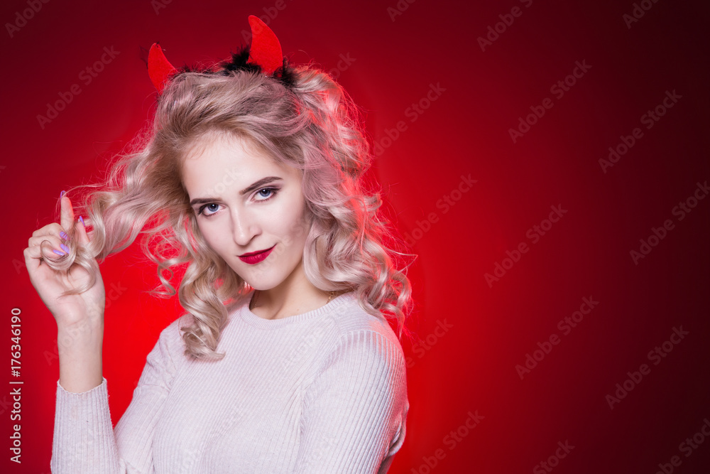Naklejka premium To twist a curl of hair on a finger. Portrait of a beautiful blonde woman with a cunning look and horns on her head. The dress for the Halloween party. Evil demon girl is the devil.