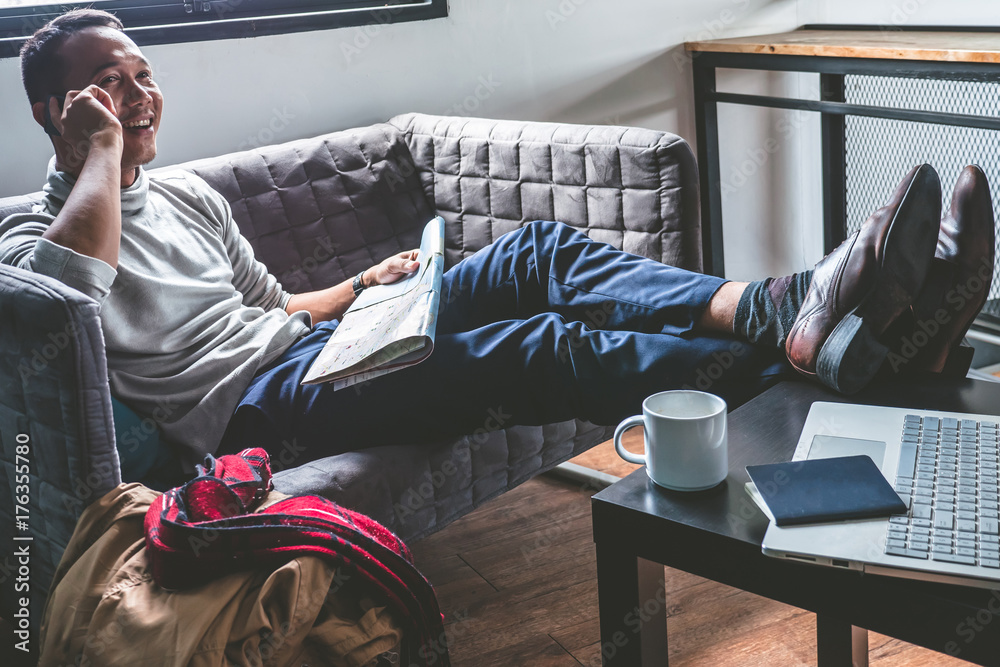 Obraz premium Asian young man using smart phone and map on sofa. Traveler man planning vacation. Travel concept.