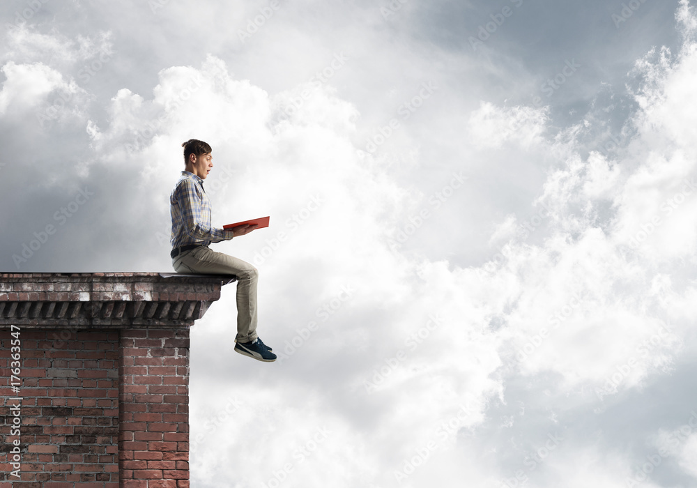 Man on roof edge reading book and cloudscape at background Man on roof edge reading book and cloudscape at background