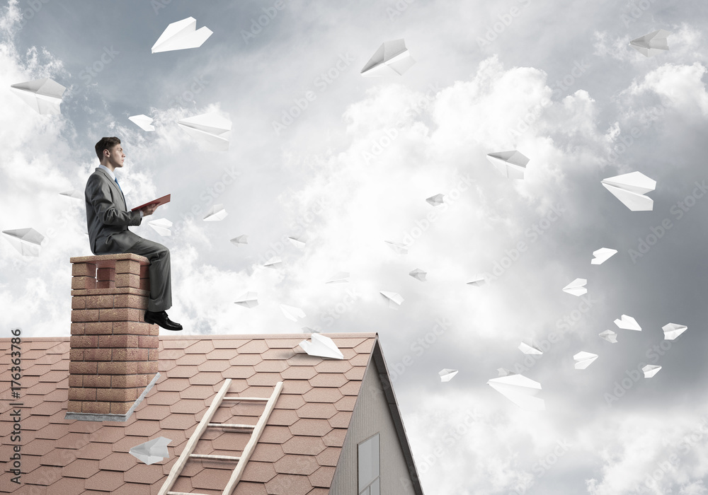 Man on brick roof reading book and paper planes flying in air Man on brick roof reading book and paper planes flying in air