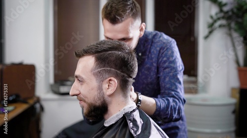 Side view of a head of stylish smiling bearded man sitting in the barbershop covered with black peignoir. Barber in casual clothes standing behind customer making boxer haircut with clipper and comb.