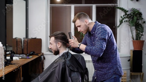 Making haircut. Young bearded man getting haircut by barber while sitting in chair at barbershop
