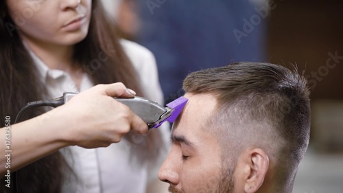 Close up of professional female hairstylist dressed in casual clothes is serving client in barbershop using modern trimmer. Young man is getting trendy haircut sitting on chair closing his eyes.