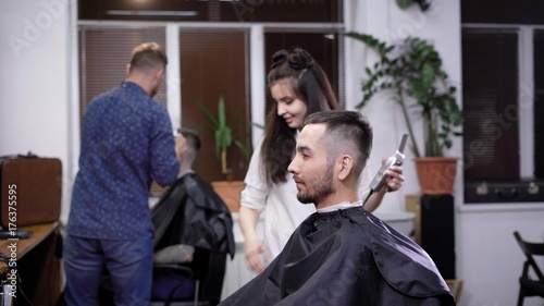 A stylish and young woman hairdresser takes a hair trimmer in her hands and begins to make hair styling and haircut for a beauty salon for a man