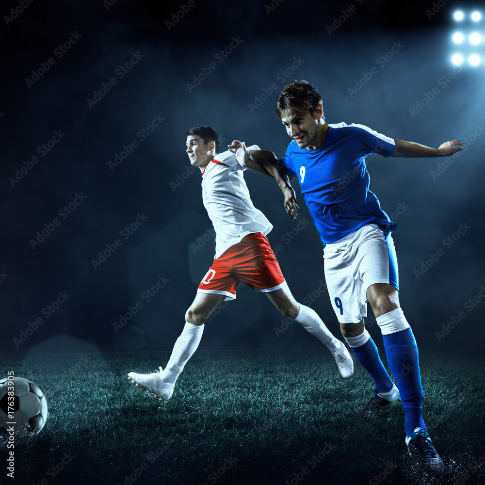 Soccer players performs an action play on a dark background. Soccer players fight for the ball. Players wears unbranded sport uniform. Soccer players performs an action play on a dark background. Soccer players fight for the ball. Players wears unbranded sport uniform.