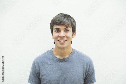 Retrato frontal de hombre con cabello castaño y camiseta gris sobre fondo blanco con expresión de  maldad.