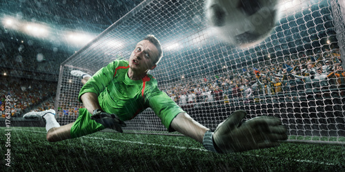 Soccer goalkeeper catches a ball on big professional rainy football arena. Goalkeeper wears unbranded sport uniform.