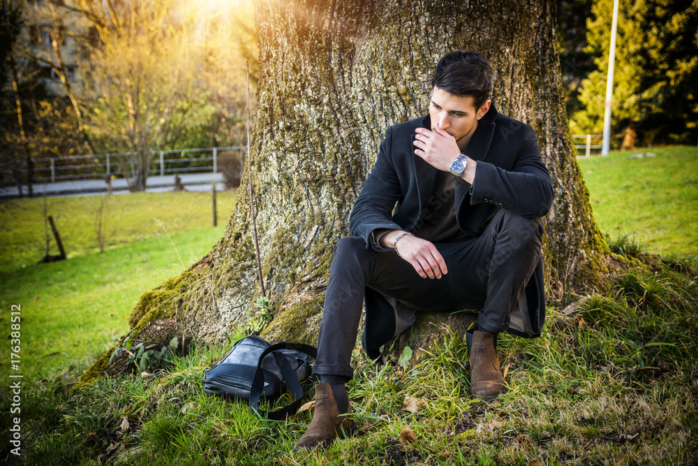 Handsome young man leaning against tree in autumn, wearing black coat Handsome young man leaning against tree in autumn, wearing black coat