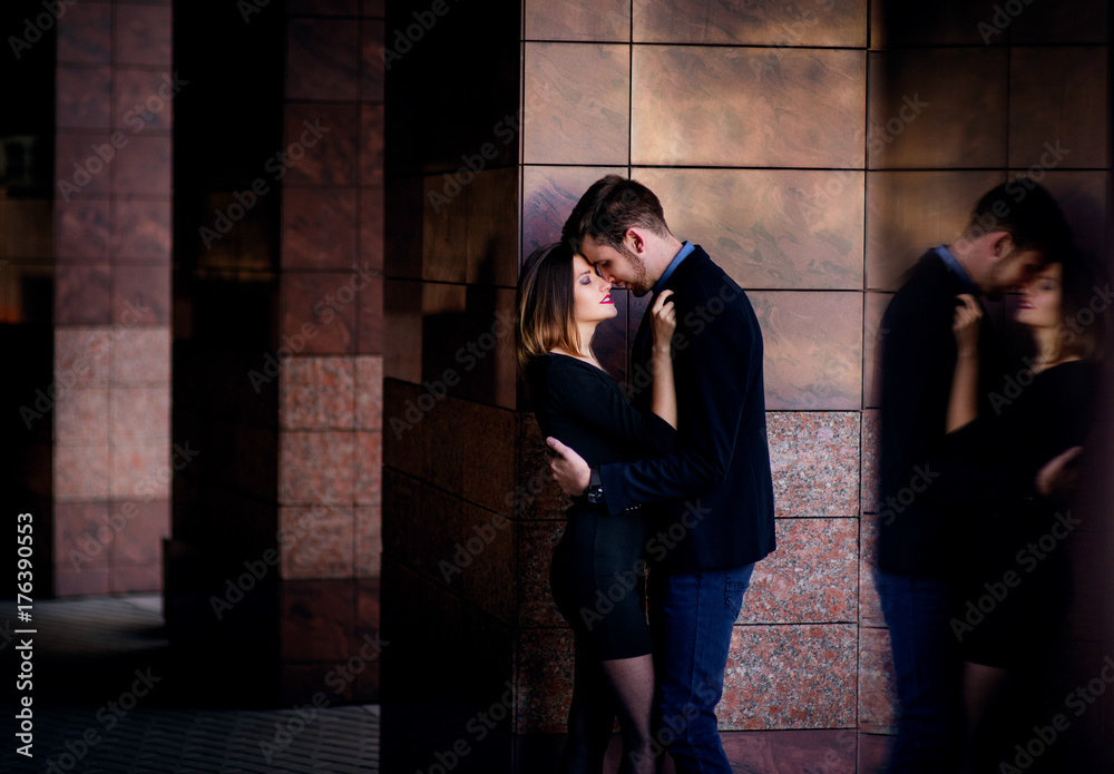 Man in black jacket and jeans leans to a marble wall hugging his girlfriend