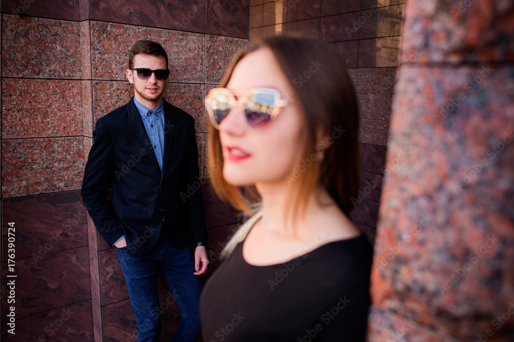 Man and woman in black sunglasses stand before marble wall