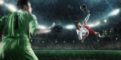 Soccer player scores the ball into the goal on professional rainy stadium. The goalkeeper protects the football gate. Players wears unbranded sport uniform.