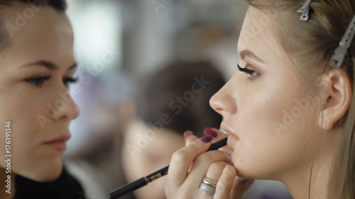 Close up of a face of young female photomodel during make-up process at beauty shop. Professional visagiste is contouring mouth line using special lip liner.