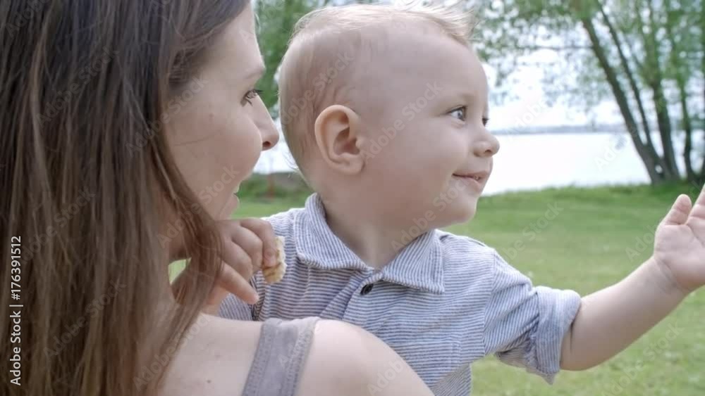 Over the shoulder shot of cheerful mother holding adorable baby boy looking at camera and laughing; green grass and lake in background Over the shoulder shot of cheerful mother holding adorable baby boy looking at camera and laughing; green grass and lake in background