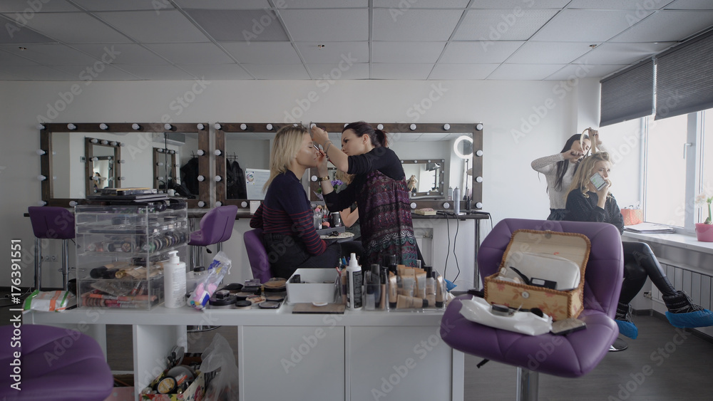 Obraz premium Beauty shop. The master in a make-up trains three girlfriends to final at university. The girl applies with a shadow brush on top. The makeup artist professionally does a make-up.