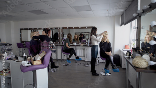 Professional hair stylist and makeup artist in their large studio preparing models before going on the podium for the show. Beautiful girl admires itself in a mirror and then looks in the smartphone.