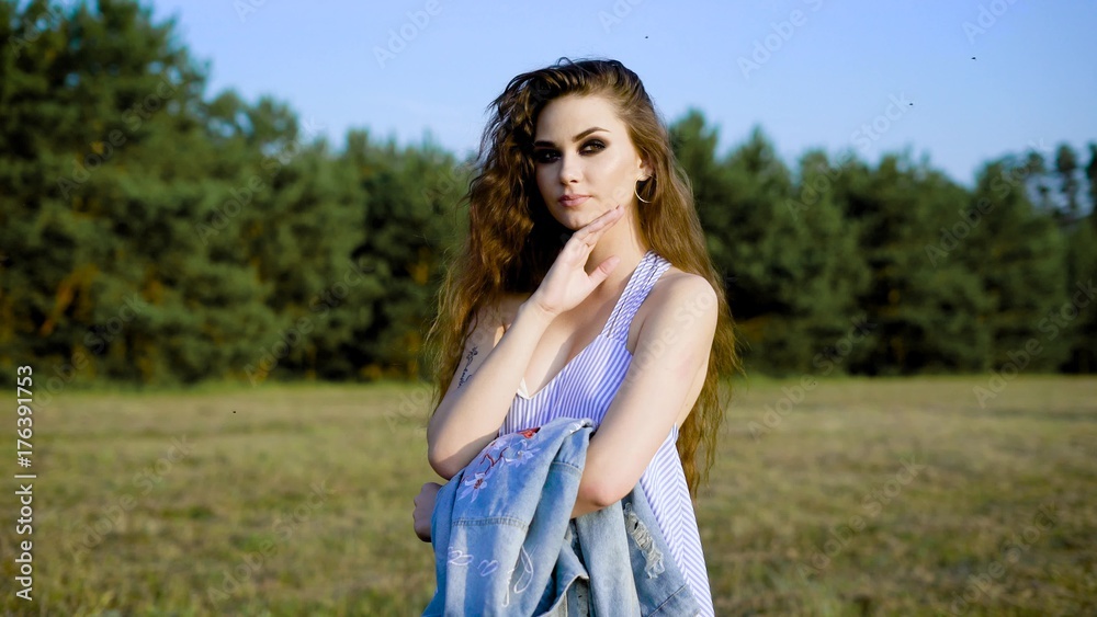 A young woman in a summer sarafan pensively stands in the nature on a summer day, a lady dresses a denim jacket on her shoulder A young woman in a summer sarafan pensively stands in the nature on a summer day, a lady dresses a denim jacket on her shoulder