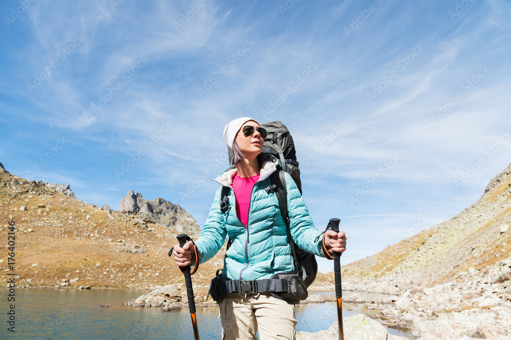 A hiker girl in sunglasses and a hat with a backpack and mountain gear with tracking treks in her hands looks at the beautiful view of a high mountain lake in an archipelago in the northern Caucasus