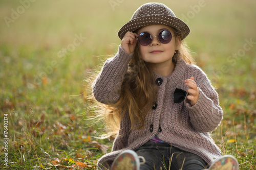 Little girl with sunglasses and hat sitting on a grass in autumn clothes