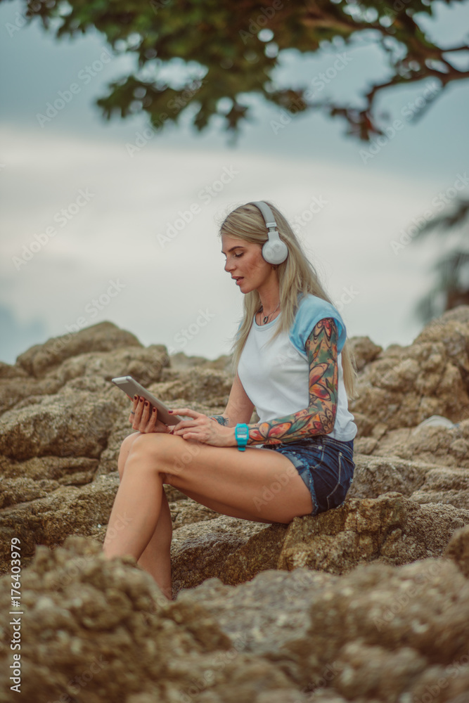 Beautiful blonde woman sitting on a rock by the sea and listening to music with your tablet. hands with tattoos. Style summer vacation. Modern girl Beautiful blonde woman sitting on a rock by the sea and listening to music with your tablet. hands with tattoos. Style summer vacation. Modern girl
