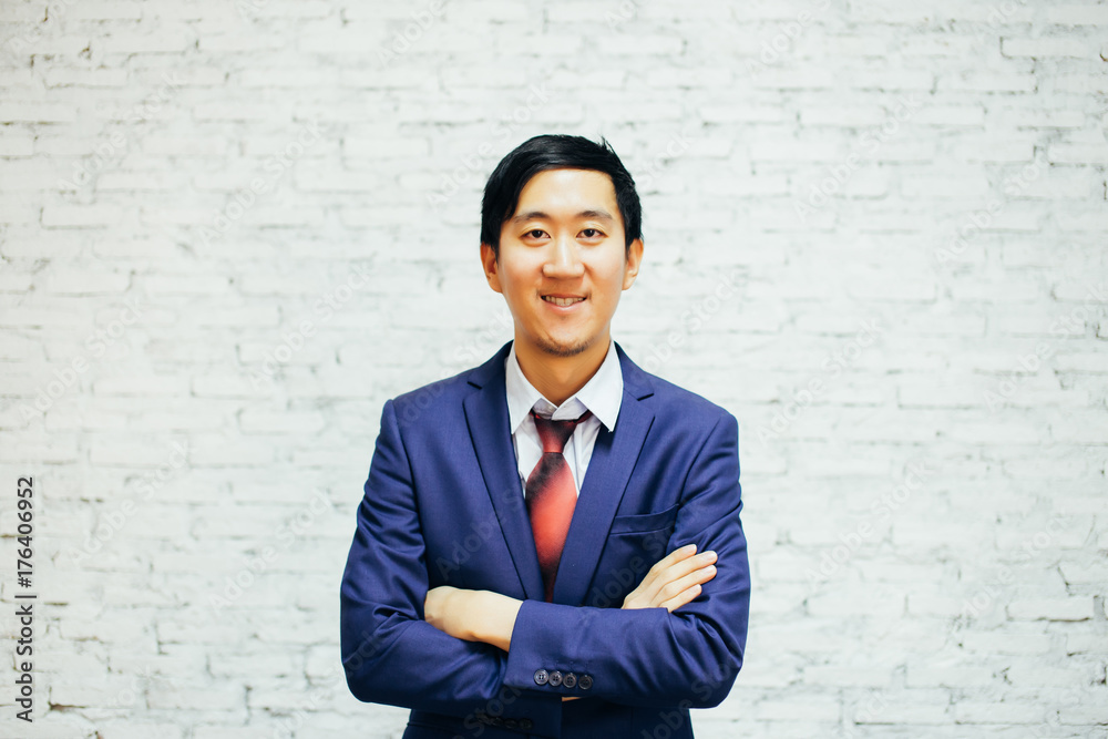 Young smiling Asian business man with arms crossed over white bricks background in the modern office - business and finance concept Young smiling Asian business man with arms crossed over white bricks background in the modern office - business and finance concept