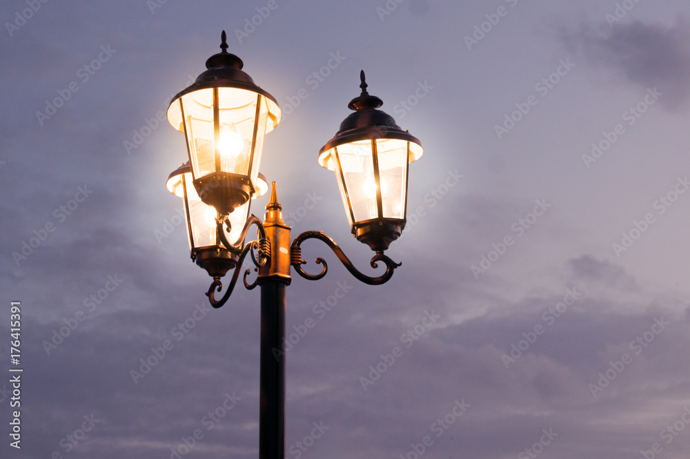 Old fashioned street lights with three lamps set on a pole shot against a cloudy sky. The dusk light and orange glow of the lamps gives this a perfect old time feel