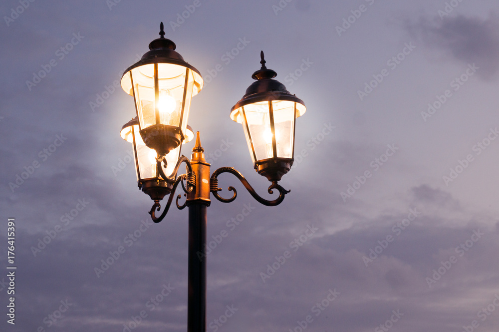 Old fashioned street lights with three lamps set on a pole shot against a cloudy sky. The dusk light and orange glow of the lamps gives this a perfect old time feel Old fashioned street lights with three lamps set on a pole shot against a cloudy sky. The dusk light and orange glow of the lamps gives this a perfect old time feel