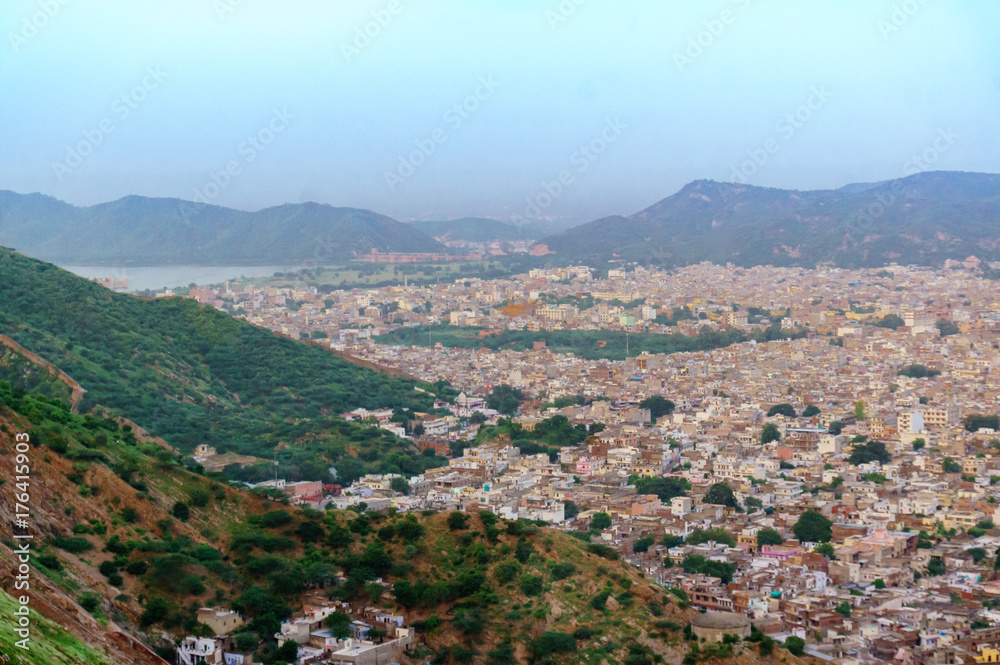 Cityscape of Jaipur city with the bordering Aravali hills and the city of Amber at the base. The cloudy sky and mountains in the background add to the beauty of the place