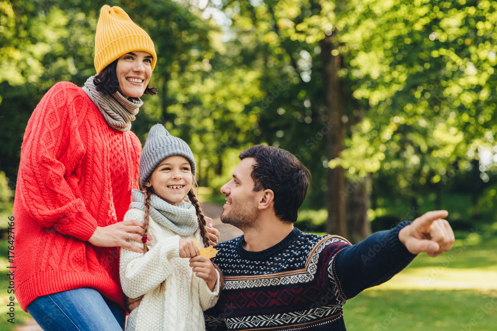 Fototapeta premium Lovely small girl with two pigtails, wears knitted hat, scarf and sweater, holds leaf in hand, looks joyfully into distance as her father shows her beautiful flower. Spending autumn days together