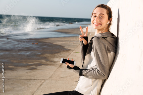 Close-up photo of happy sport woman showing peace sign, looking at camera while listening to music