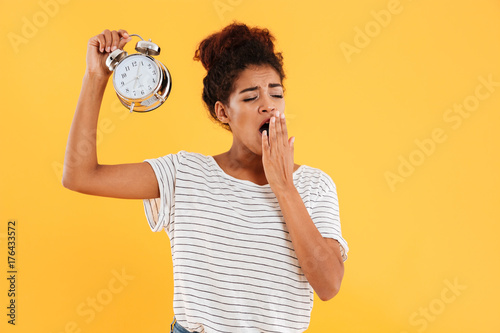 Tired african woman yawns and holding alarm clock