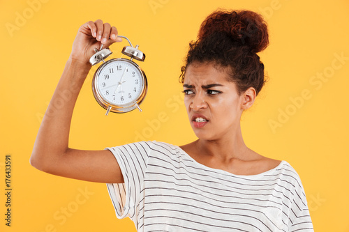 Displeased african woman holding alarm clock and looking on them