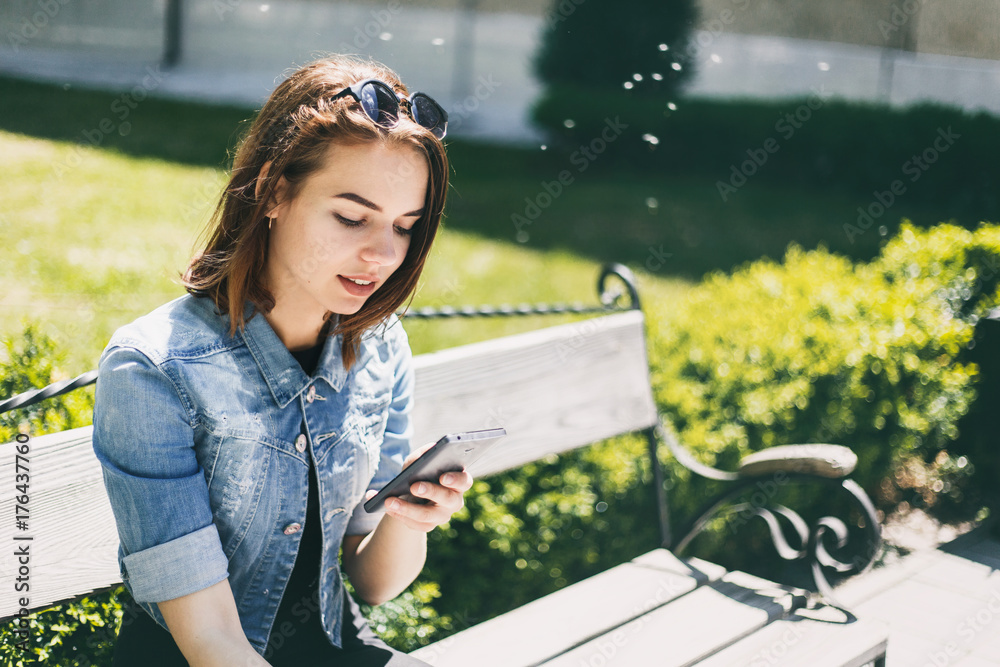 Naklejka premium Beautiful cheerful girl with a smartphone sitting in a park on a bench