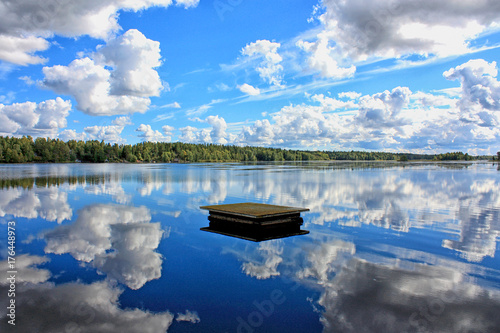 Tranquil landscape at the lake, with the blue sky, white clouds and a forest reflected symmetrically in the clean blue water
