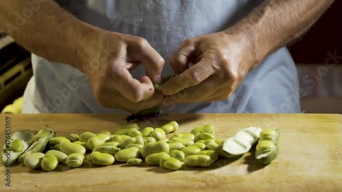 Wide, slider shot from right to left of a cutting board with shelled fava beans on the board and a man opening the pod with his fingers and pulling out the fava beans. photo