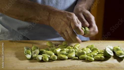 Wide, slider shot from left to right of a cutting board with shelled fava beans on the board and a man in a blue shirt opening the pod with his fingers and pulling out the fava beans. photo