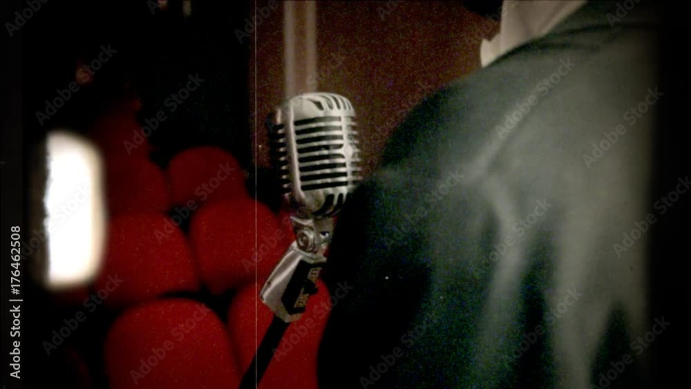 Fake 8mm amateur film: a singer in front of a retro vintage shiny microphone on the stage of a theater or cinema. Red seats in the background.
