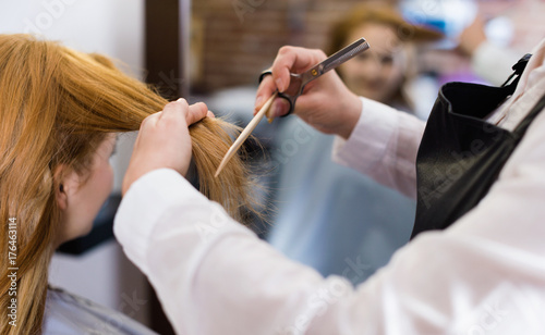 Hairdresser leveling and cutting hair to young blonde woman with use of scissors