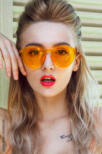 Close up portrait of a beautiful young blonde girl with a stylish orange sunglasses that fits well with her orange bikini and red lipstick. Background for this image are wooden slatted blinds.
