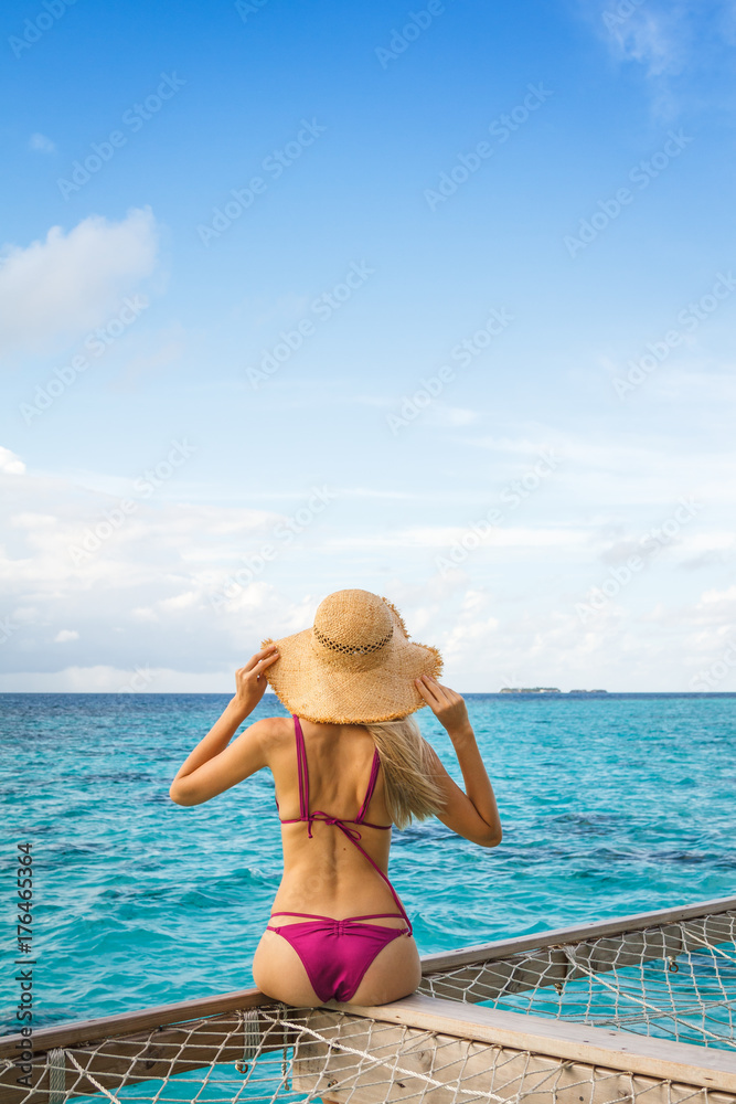 A beautiful young blonde woman sitting on a terrace above the open ocean. Tans in a purple bikini and a big straw hat. Enjoying the fresh air and the wind. Interesting terrace is made of wood and net.