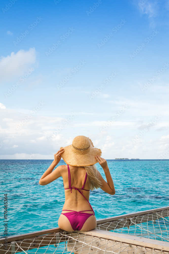 A beautiful young blonde woman sitting on a terrace above the open ocean. Tans in a purple bikini and a big straw hat. Enjoying the fresh air and the wind. Interesting terrace is made of wood and net. A beautiful young blonde woman sitting on a terrace above the open ocean. Tans in a purple bikini and a big straw hat. Enjoying the fresh air and the wind. Interesting terrace is made of wood and net.