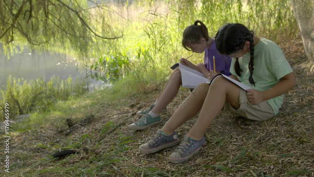 Two girls sit on ground under willow tree next to pond and draw in notebooks during an outdoor class at summer camp. Hand-held slow motion 4K recorded at 60fps.