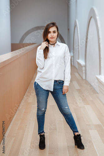 Portrait of sexy white Caucasian brunette young beautiful girl woman with  long dark hair in white shirt and blue jeans. Young girl standing indoor in  hall at college university. Stock Photo