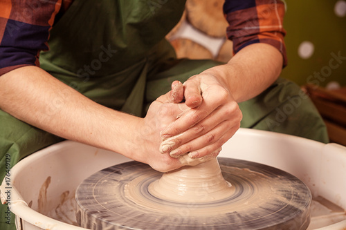  Young woman potter in a green apron and checkered shirt beautifully and smoothly sculpts a brown clay vase on a potter's wheel, side view