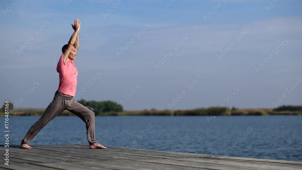 A man wearing pink shirt and grey yoga pants doing yoga, warrior position, outdoor in the quiet scenery by the lake / A man doing yoga by the lake A man wearing pink shirt and grey yoga pants doing yoga, warrior position, outdoor in the quiet scenery by the lake / A man doing yoga by the lake