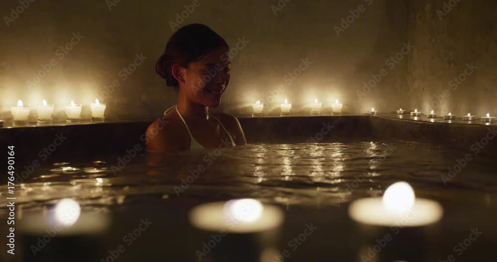 Beautiful woman entering Jacuzzi. Happy female is relaxing in hot bath tub. Beauty spa is decorated with lit candles enjoying vacations.