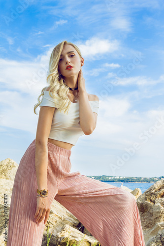 Beautiful young woman with long curly blonde hair with a beautiful makeup posing on the harbor. In the background are beautiful clouds. Around her neck is a stylish black choker.