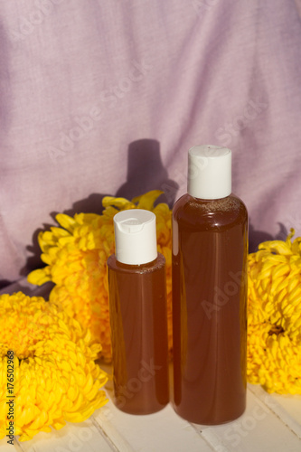 Two bottles with gel stand on white wooden table in front of yellow chrysanthemums and purple fabric.