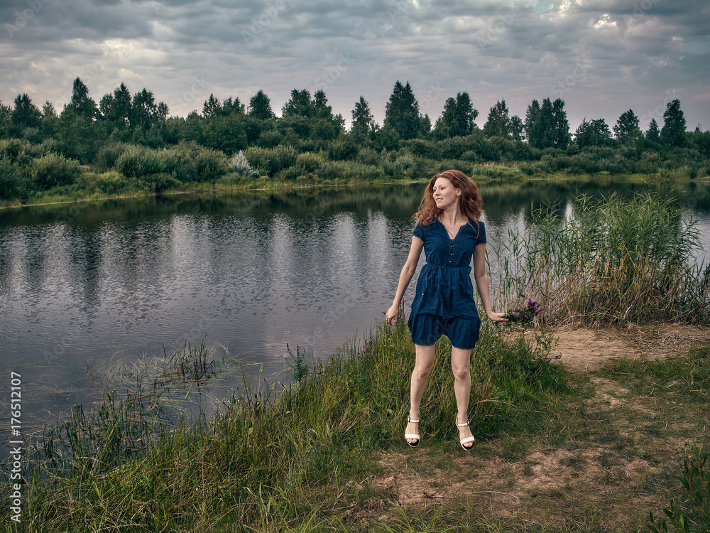 The red-haired girl in a blue summer dress strolls along the lake.