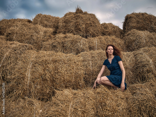Red-haired girl in a light summer dress of blue color sits on a stack of straw.